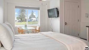 Une chambre à coucher comprenant un lit, un bureau, une chaise, un téléviseur et une fenêtre donnant sur des arbres et des maisons.