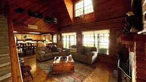 A cozy cabin interior with a wooden ceiling, a fireplace, a dining area, and two couches.