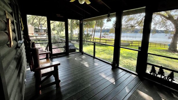 A covered porch with a ceiling fan, wooden flooring, and large windows offering a view of a lake and grassy area.