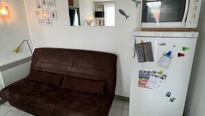 Habitació petita i senzilla amb un sofà marró, un refrigerador blanc amb imants i notes, un microones i una petita decoració de peix a la paret.