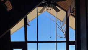Ein gemütliches Zimmer mit großem Fenster, Deckenventilator aus Holz und Blick auf eine Terrasse und die Landschaft.