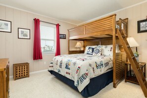 A bunk bed with a nautical-themed bedspread, a window with red curtains, and a small wooden side table with a lamp.