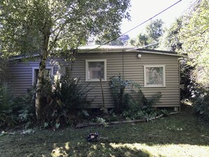 Une maison de plain-pied avec un porche à l’avant, entourée d’une végétation luxuriante et d’un arbre.