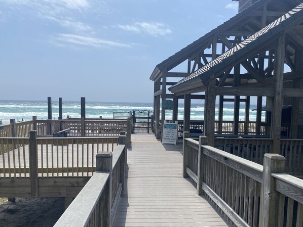 Une promenade en bois mène à une zone de plage où un panneau indique qu’il est nécessaire de s’enregistrer au bar pour recevoir un bracelet.