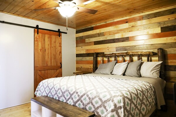 A bedroom with a wooden slat wall, a bed with patterned bedding, a wooden bench, and a ceiling fan.