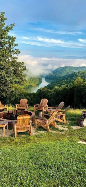 A circular wooden seating arrangement with a fire pit, overlooking a river and forested hills.