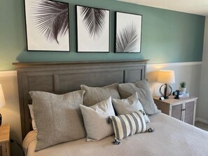Una cama con varias almohadas, una mesita de noche con lámpara y tres cuadros con hojas de palma.