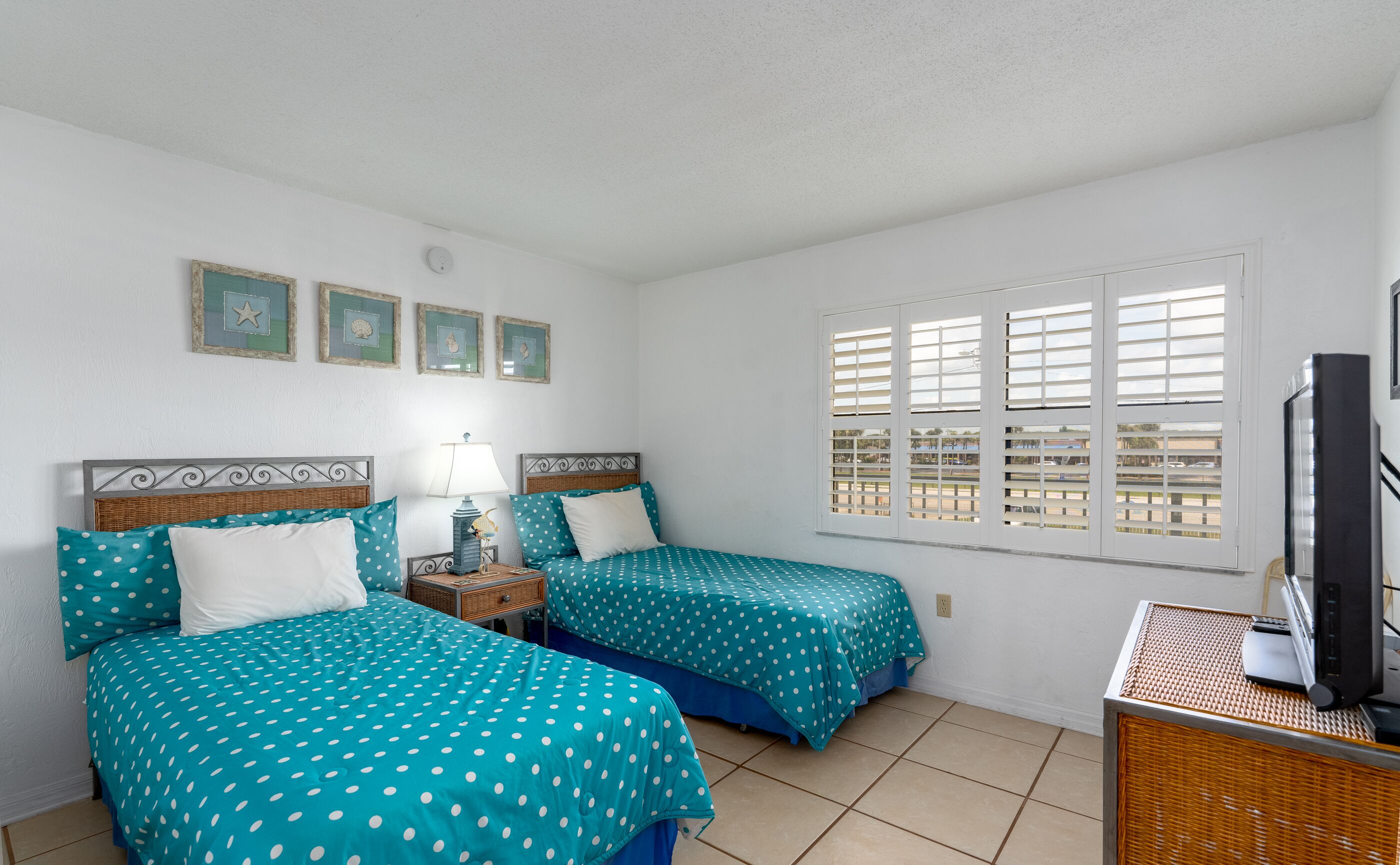 A room with two beds, a window with blinds, a lamp, and a television on a stand.