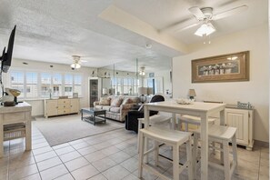 Un salon spacieux comprenant un coin repas, de grandes fenêtres, des ventilateurs de plafond et un tableau encadré accroché au mur.