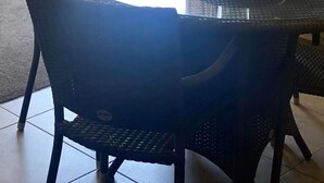 Una mesa redonda de vidrio con un florero y un cuenco, rodeada de sillas de mimbre, en una habitación con vistas a la vegetación exterior.