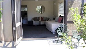 Un dormitorio con cama, un sofá y una pequeña mesa con sillas. Hay una ventana con persianas y una puerta que da a otra habitación.