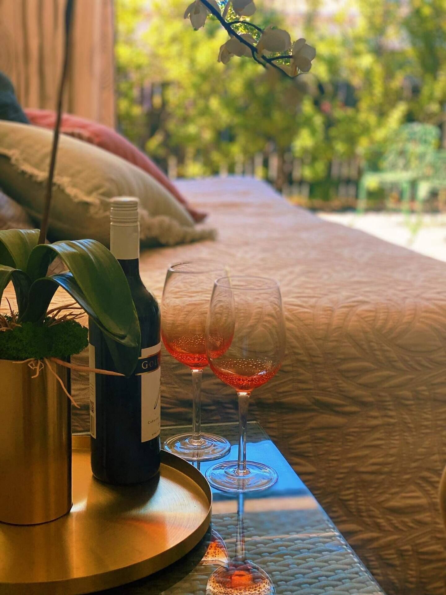 En säng med utsikt över grönskan, en flaska vin och två glas på ett brickbord.
