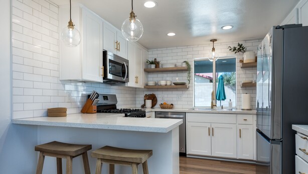 Uma cozinha moderna com armários brancos, uma ilha central com banquetas, micro-ondas e frigorífico.