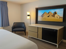 Habitación de hotel con cama, escritorio con cajones, silla, lámpara y televisor montado en la pared mostrando un paisaje desértico.