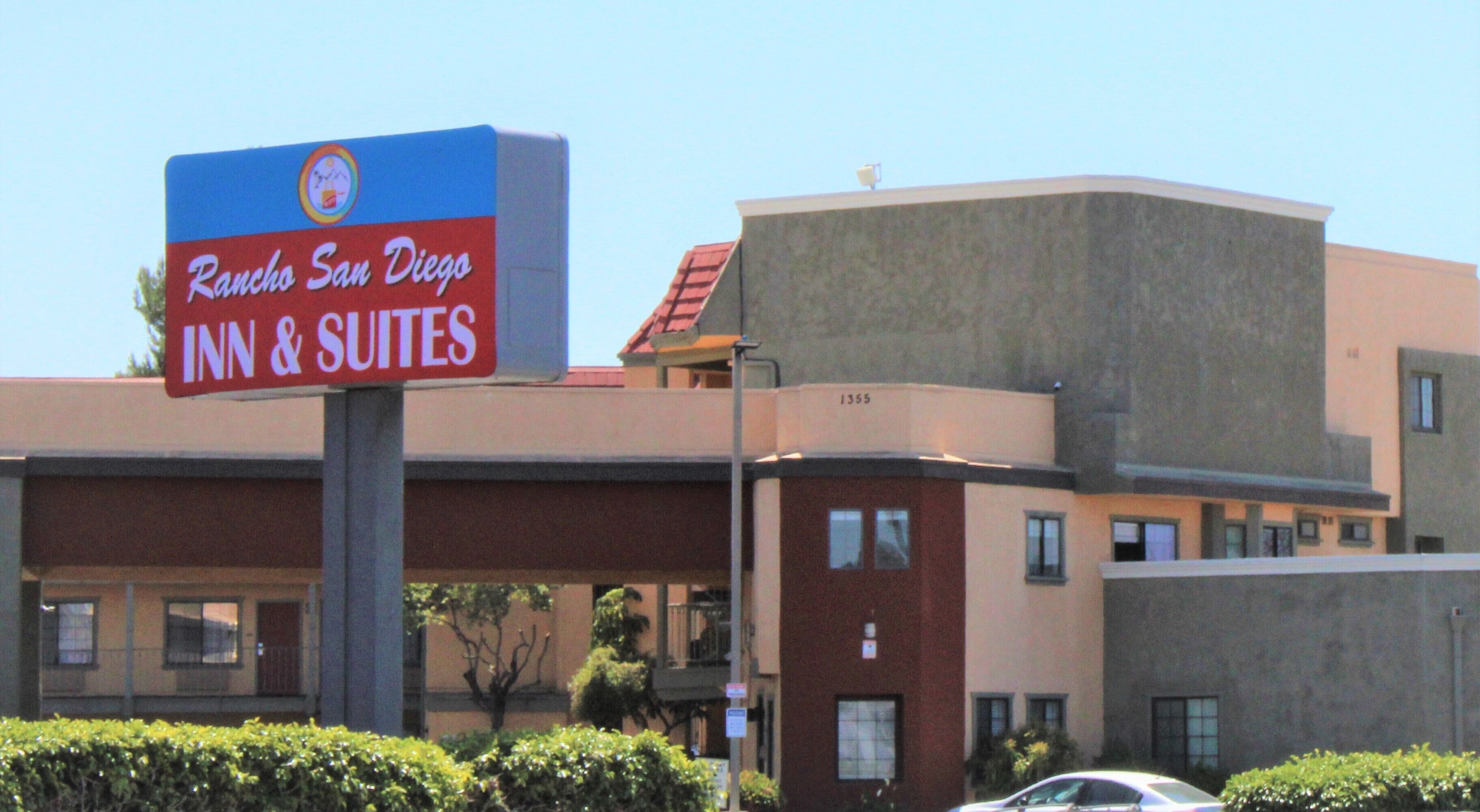 A sign for Rancho San Diego Inn & Suites, featuring a modern building with a red roof and a number '1355' on the side.