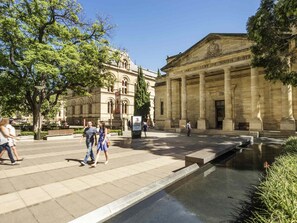Una plaza pública con una fuente, un gran edificio de estilo clásico con columnas, y gente caminando.