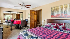 Habitación de hotel con una cama grande, un ventilador de techo, un comedor con mantel rojo y un cuadro de un bosque.