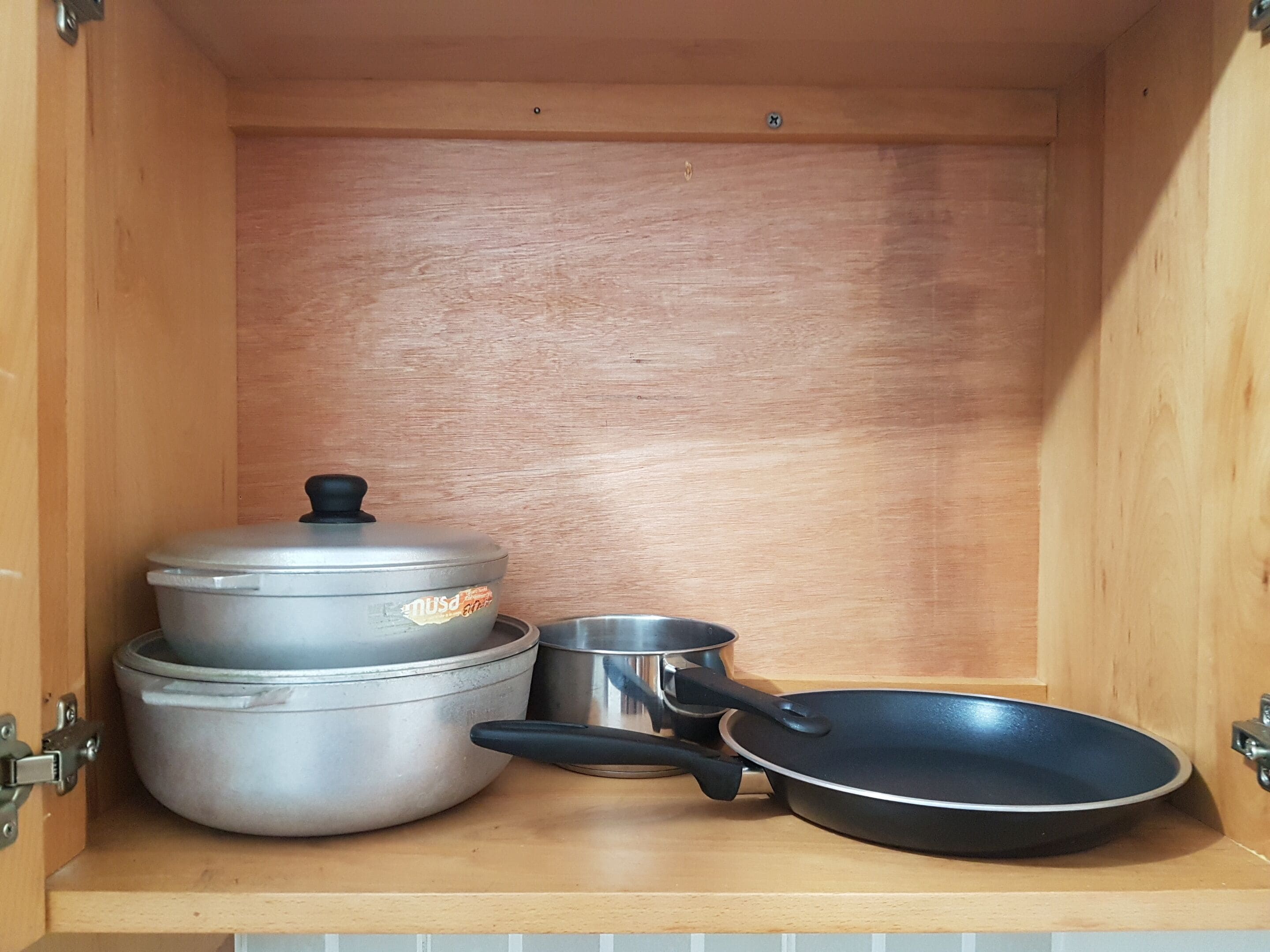 A kitchen cabinet with a wooden interior, containing a stack of pots and pans, including a large pot with a lid, a smaller pot, and a frying pan.