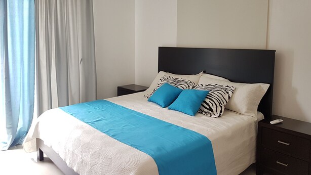 A neatly arranged bedroom with a bed, nightstands, a ceiling fan, and a window with curtains. - Céntrico Próximo Clínica Unión Médica (Santiago de los Caballeros)