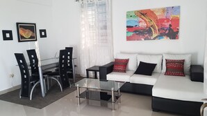 A modern living room with a glass coffee table, a white sofa with patterned cushions, a dining area with black chairs, and a large abstract painting on the wall. - Céntrico Próximo Clínica Unión Médica (Santiago de los Caballeros)