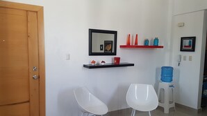 A clean, modern room with a wooden door, white walls, a black-framed mirror, a red shelf with decorative items, two white chairs, and a water dispenser. - Céntrico Próximo Clínica Unión Médica (Santiago de los Caballeros)