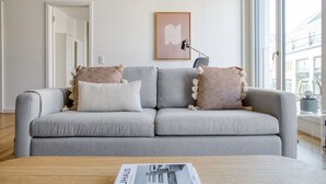 A modern living room with a grey sofa, two side tables, a framed picture, and a window with a view of buildings. - Gorgeous Mitte 1BR with Apple Store in building, by Blueground (Berlin)