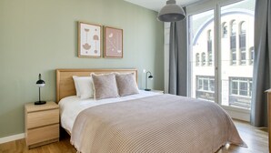 A bedroom with a wooden headboard, a large bed with a textured blanket, two bedside tables with lamps, and a view of a building through a window. - Gorgeous Mitte 1BR with Apple Store in building, by Blueground (Berlin)