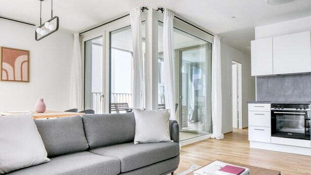 A modern living room with a grey sofa, a wooden coffee table, and a kitchen area with a built-in oven. - Deluxe Leopoldstadt 2BR w/ Loggia, Gym & Sauna, near U2, by Blueground (Vienna)