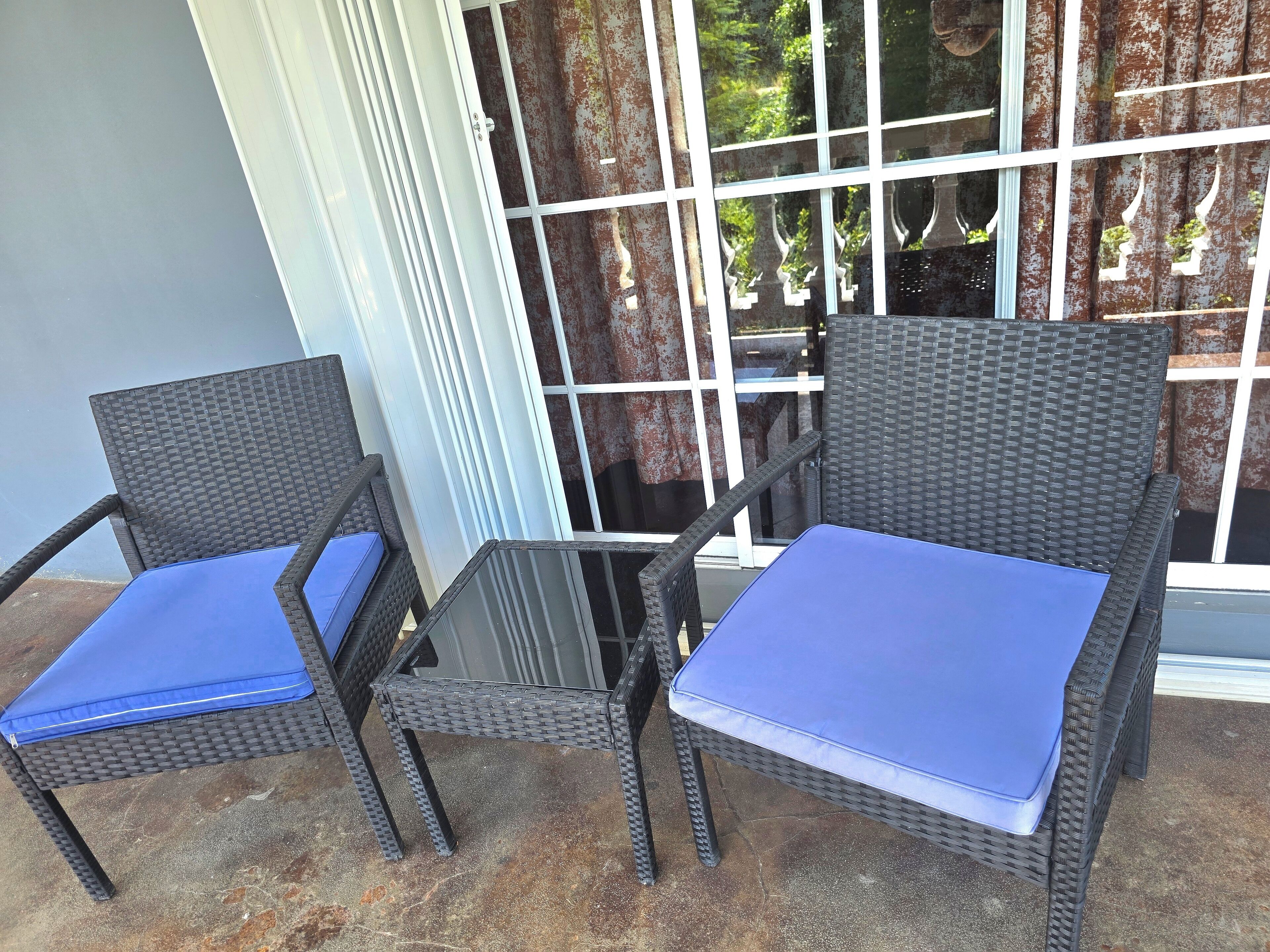 Zwei Korbstühle mit blauen Kissen und ein Glastisch stehen auf einer Terrasse, von der aus ein Gebäude durch ein Fenster mit Gitterrost sichtbar ist.