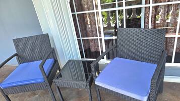 Zwei Korbstühle mit blauen Kissen und ein Glastisch stehen auf einer Terrasse, von der aus ein Gebäude durch ein Fenster mit Gitterrost sichtbar ist.