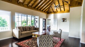 A spacious living room with a high ceiling, large windows, a sofa, patterned chairs, and a wooden coffee table. - Blue Paloma Bed & Breakfast (Lucea)