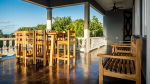 A covered outdoor area with wooden furniture, including chairs and a table, and a view of lush greenery and a clear sky. - Blue Paloma Bed & Breakfast (Lucea)