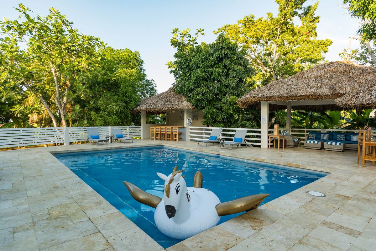 Ein Swimmingpool mit einem Schwanschwimmpfad, Liegestühlen und strohgedeckten Gebäuden.