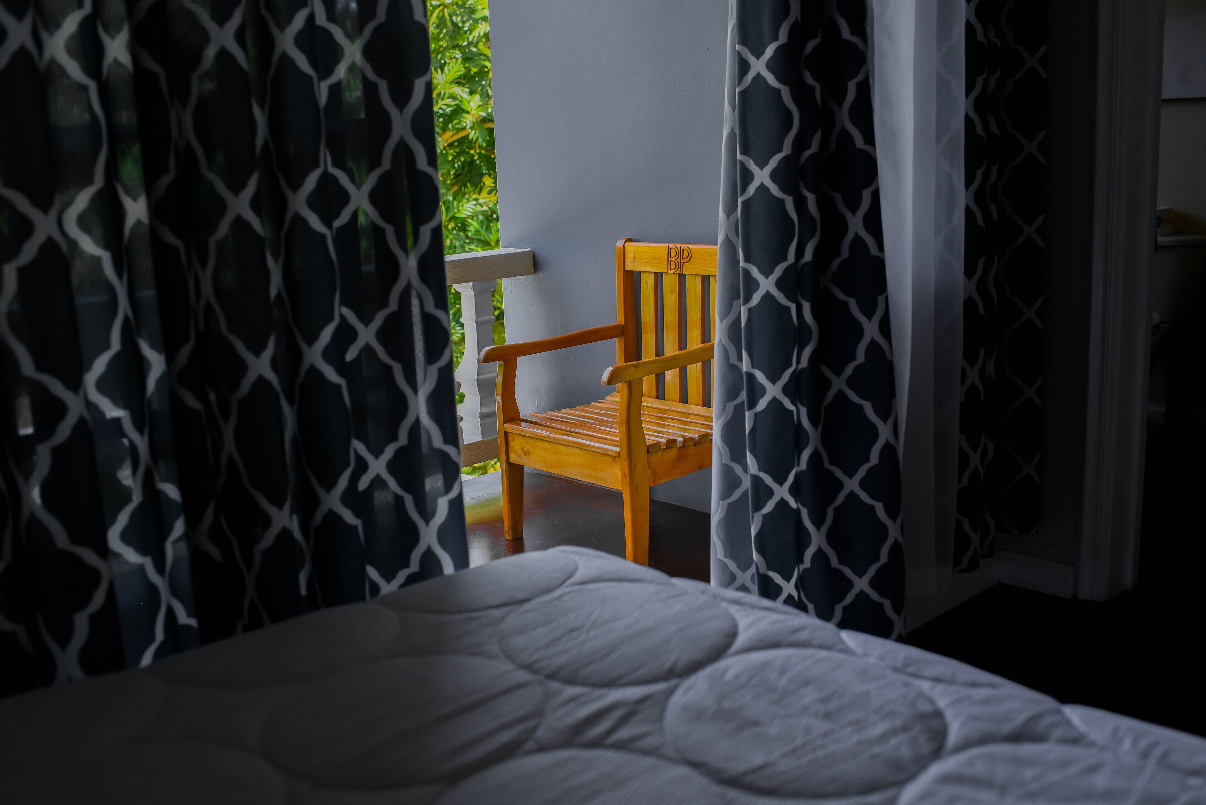 Una cama con colcha blanca, una silla de madera con reposacabezas y cortinas estampadas.