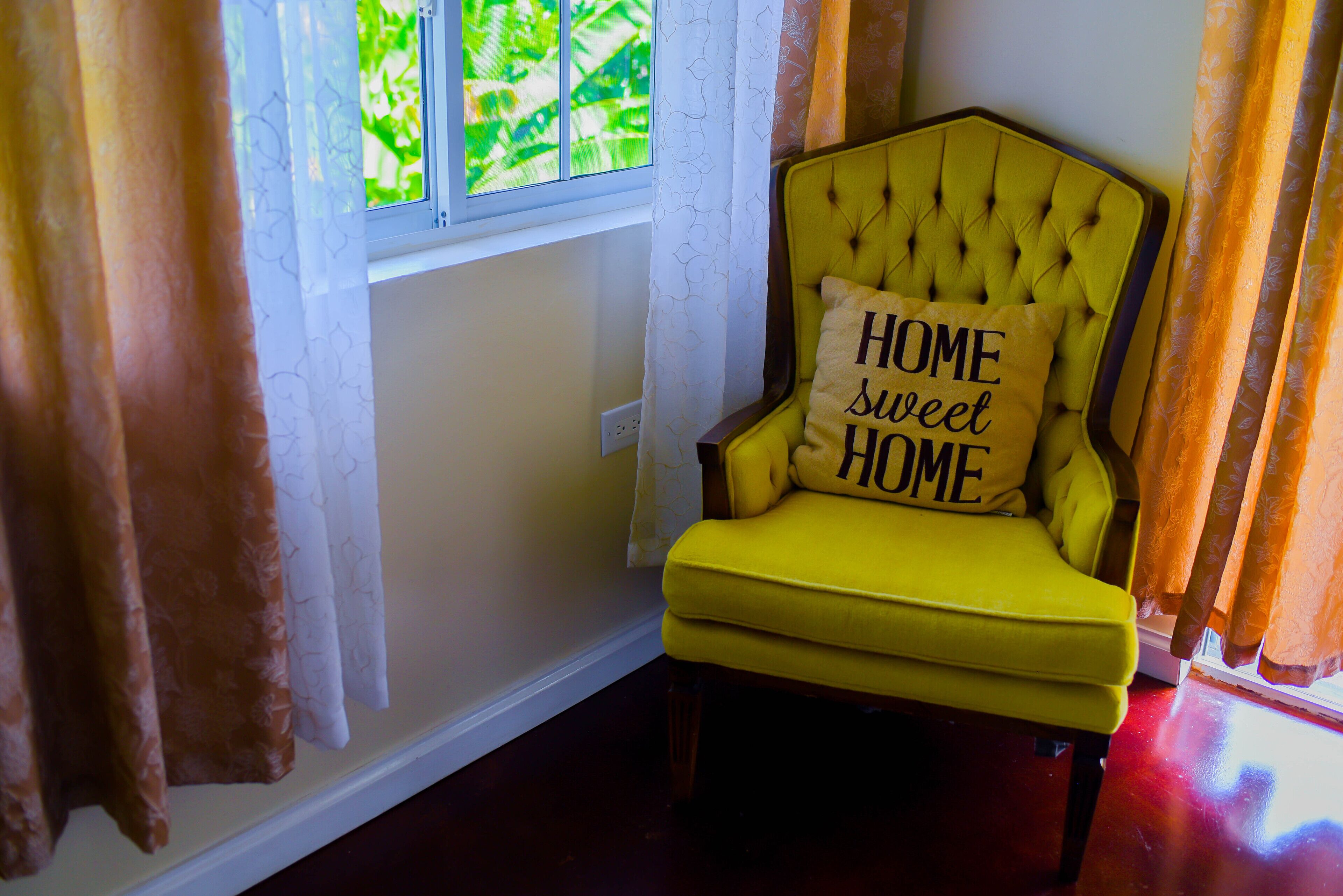 Un sillón amarillo con un cojín que dice 'HOME sweet HOME' junto a una ventana con cortinas estampadas.