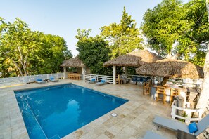 A swimming pool with lounge chairs and thatched umbrellas, surrounded by trees and a white fence. - Blue Paloma Bed & Breakfast (Lucea)
