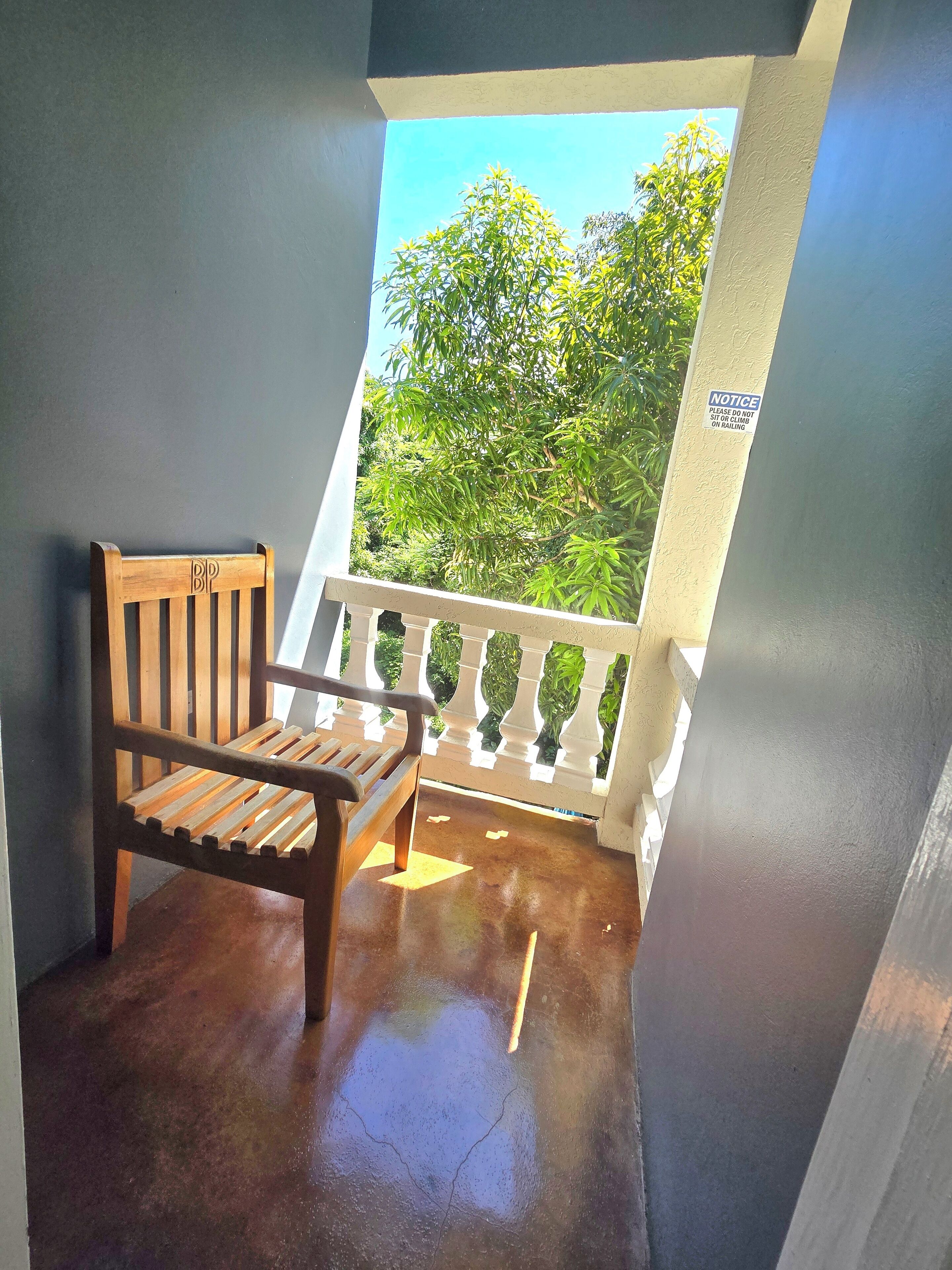A wooden chair on a balcony with a white railing, a notice sign, and a view of trees.