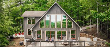 Une cabane à deux étages dotée de grandes fenêtres, d’une terrasse et d’un espace salon extérieur.