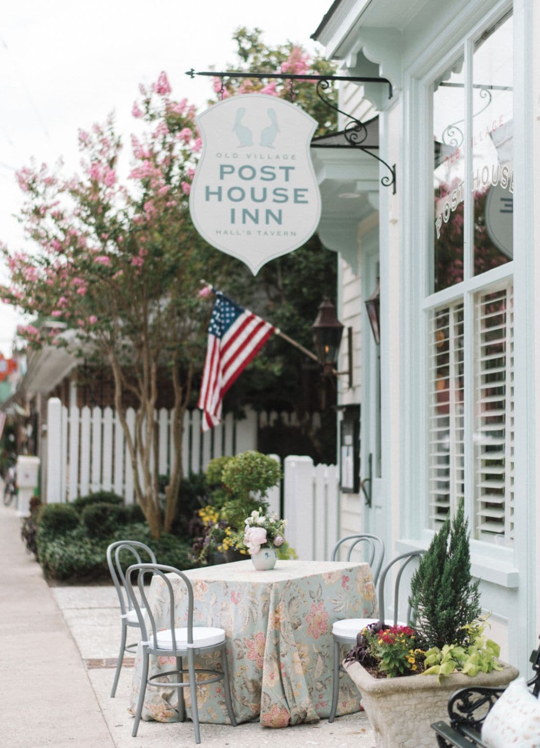 Une enseigne indiquant « Post House Inn », des tables et des chaises disposées à l’extérieur, et une clôture en bois peinte en blanc en arrière-plan.