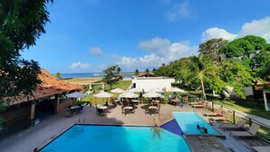 A pool area with lounge chairs and umbrellas, a view of the ocean, and a building in the background. - Catamaran Praia Chalés (Igarassu)