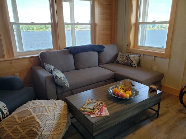 A cozy living room with a brown sofa, a wooden coffee table with magazines and fruit, and large windows overlooking a body of water.