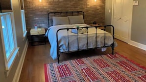 Een slaapkamer met een bakstenen muur, een metalen bedframe, een houten nachtkastje, een rood tapijt met een patroon en een witte deur.