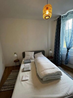 Una cama bien tendida con almohadas y toallas dobladas, un colgante de luz, una mesita de noche con lámpara y una ventana con cortinas.