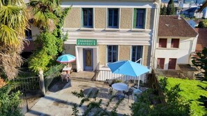 Un edificio a due piani con un cartello che indica “CHAMPRES ENTRÉE”. Sono presenti tavoli e ombrelloni all'aperto, e una scala che conduce all'ingresso.