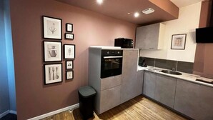 Eine moderne Küche mit grauen Schränken, einem eingebauten Backofen und einer Mikrowelle darüber. An der Wand hängen Bilder in Rahmen, und ein schwarzer Mülleimer steht bereit.