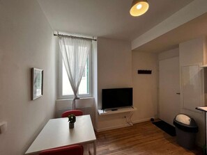 A small, well-lit room with a wooden floor, a white table, a red chair, a TV mounted on the wall, and a window with sheer curtains.