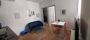 A compact living space with a sofa, a dining table with chairs, a small stool, and a refrigerator.