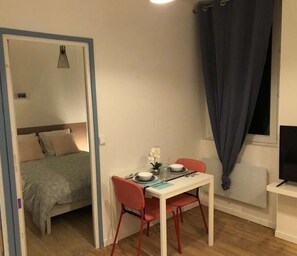 A compact room with a bed, a dining table with chairs, a small table with a vase of flowers, and a TV on a stand.
