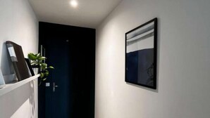 Um corredor estreito com piso de madeira, uma porta escura no final, um quadro na parede e uma planta à esquerda.