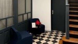 Une pièce avec un sol à carreaux, une chaise noire, une petite table sur laquelle se trouve un objet rouge, et une porte.